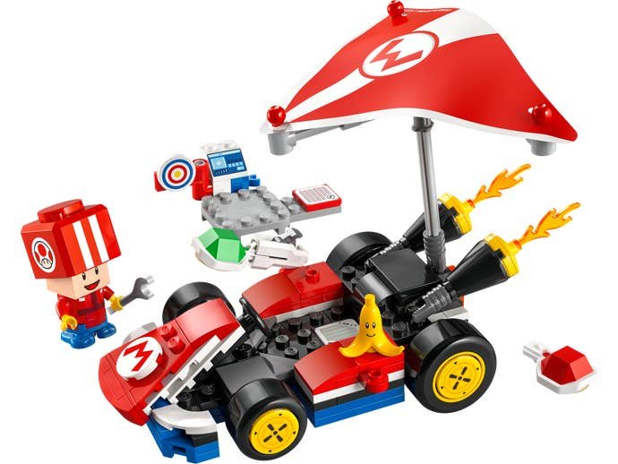 A red kart from Mario, equipped with a paraglider, and attended by a small Toad mushroom creature. A red shell is behind the vehicle.